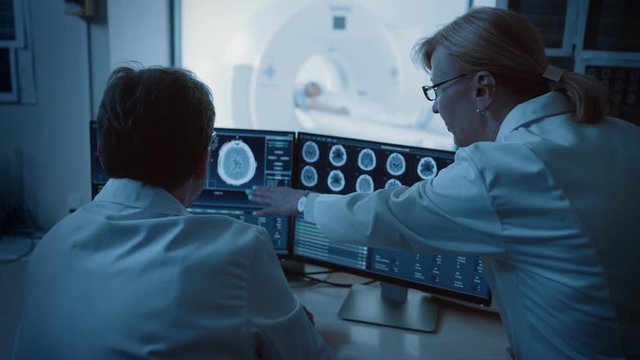 In Control Room Doctor and Radiologist Discuss Diagnosis while Watching Procedure and Monitors Showing Brain Scans Results, In the Background Patient Undergoes MRI or CT Scan Procedure.