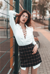 Young fashion girl with red hair dressed in a plaid skirt and white shirt posing in a park near the sports field. Street style autumn.