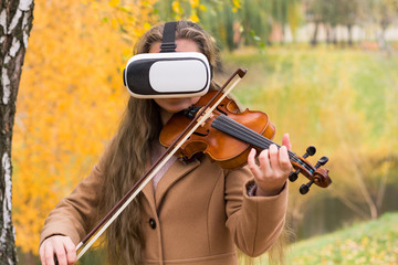 Girl playing the violin in virtual reality glasses in the autumn park at a lake background.