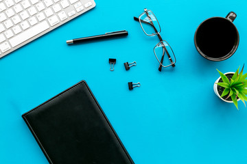 Office desk of creative person or hipster.Cute workplace. Keyboard and glasses near coffee, notebook, green room plant and stationery on blue, turquoise background top view copy space