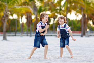 Fototapeta na wymiar Two little kids boys having fun on tropical beach, happy best friends playing, friendship concept. Siblings brothers, twins in family look with palms trees on background. Family vacations.