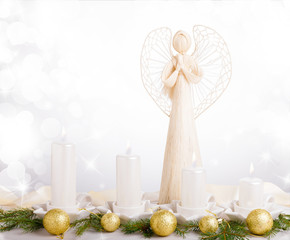 A white angel and four white Advent candles, Christmas tree branches are decorated with golden...