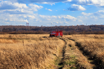 Fototapeta premium Red firetruck car Ural rides through the autumn field with yellow and faded grass against the blue sky and clouds. The concept of extinguishing forest fires in USA