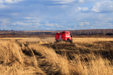 Fototapeta premium Red firetruck car Ural rides through the autumn field with yellow and faded grass against the blue sky and clouds. The concept of extinguishing forest fires in USA