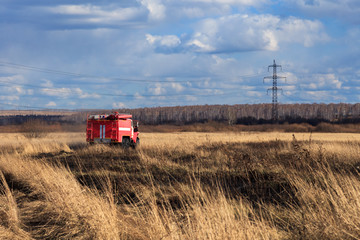 Fototapeta premium Red firetruck car Ural rides through the autumn field with yellow and faded grass against the blue sky and clouds. The concept of extinguishing forest fires in USA
