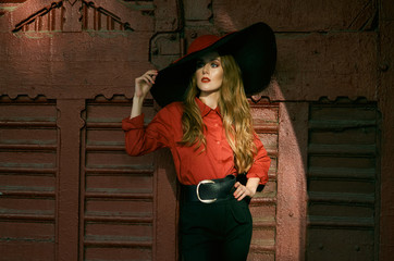 Attractive slim woman in a wide-brimmed hat, a red blouse and black pants posing against a vintage red door. Fashionable photo.