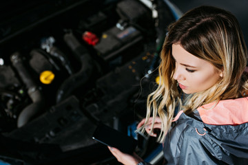 Plakat high angle view of young woman using smartphone with blank screen while repairing broken car