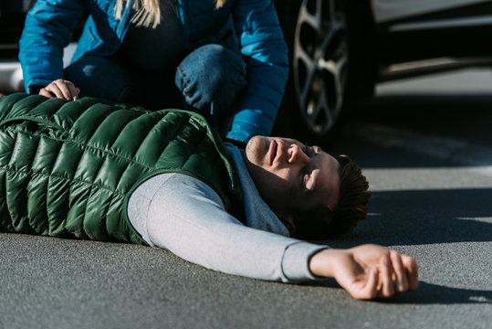 Cropped Shot Of Woman Touching Injured Young Man Lying On Road After Car Accident