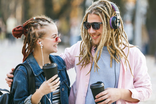 Two hipster best friends with dreads wearing headphones and glasses laughing and drinking coffee while embraicing and looking for each other in the street