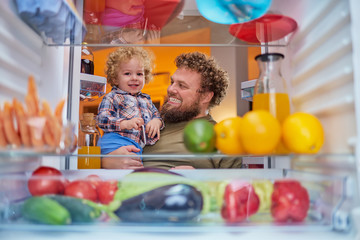 Fototapeta na wymiar Father and son standing in front of opened fridge and looking something to eat at night. Fridge full of groceries. Picture taken from the inside of fridge.