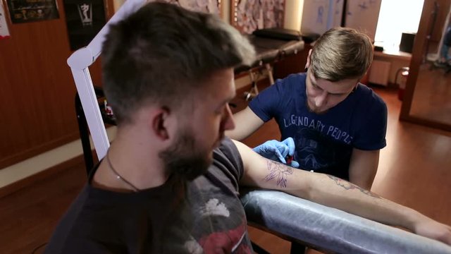 Master doing tattooing in tattoo studio. Professional tattooist works in studio. A man in blue gloves makes a tattoo on the hand of a young guy.