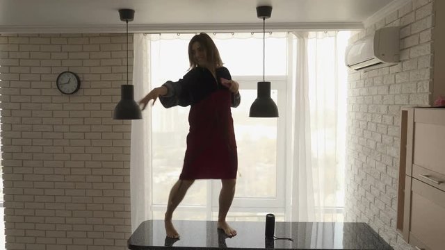 A pretty brunette girl in a red velvet dress is dancing barefoot on the black kitchen table with a cellphone in her hand and a wireless speaker on the table