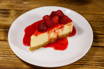 Piece of tasty New York cheesecake with raspberries and raspberry jam in a white plate on wooden table