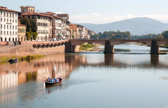 Riverboat with tourists floating past river bridge of ancient Tuscany city