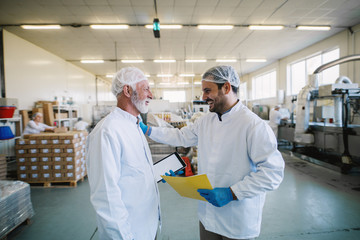 Two cheerful male food factory employees in sterile clothes smiling and talking about business plans.