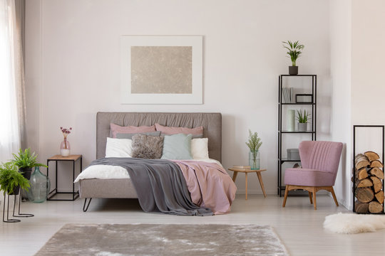 Pink and grey sheets on bed next to armchair in bedroom interior with plants and poster. Real photo