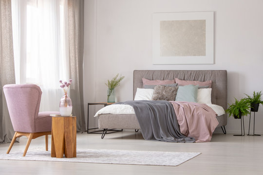 Flowers on wooden table next to pink armchair in grey bedroom interior with poster above bed. Real photo