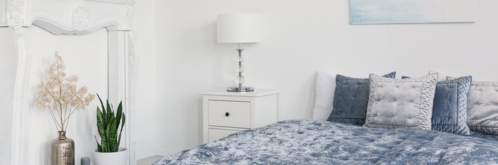 Panorama of plants and lamp on cabinet in white bedroom interior with cushions on blue bed. Real photo