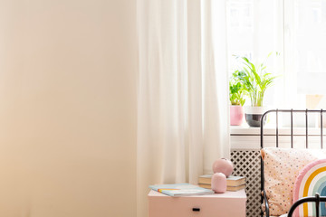 Copy space on white wall in girl's bedroom interior with plants and pink cabinet next to bed. Real...