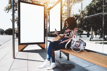 A cheerful young caucasian woman with bulky curly hair is sitting on a yellow metal bench inside of an urban bus stop and using her cell phone, with two white blank info banner templates behind her - obrazy, fototapety, plakaty