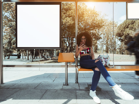 A smiling caucasian female with bulky curly hair and in eyeglasses is sitting inside of the outdoor bus stop and using her smartphone with an empty white mockup of information billboard next to her