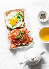 Served breakfast - toast with cream cheese, fried egg, tomatoes, bacon and green tea on a light background, top view