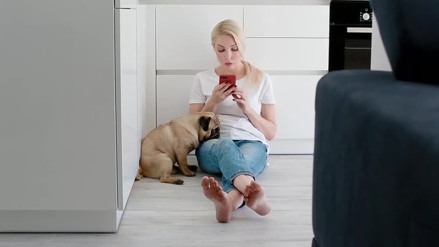Woman pet the dog while using smartphone on the kitchen. Pug sitting next to the owner.