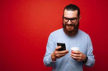 Happy bearded hipster man in casual using phone and hoding cup of coffee over red background