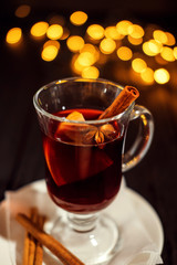Fototapeta premium closeup glass of mulled wine with orange and cinnamon on dark black background, on white plate, Christmas lights