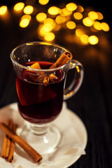 closeup glass of mulled wine with orange and cinnamon on dark black background, on white plate, Christmas lights, large yellow bokeh