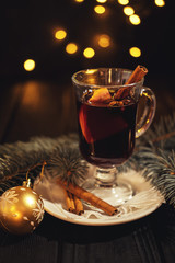 closeup glass of mulled wine with orange and cinnamon on dark black background, on white plate, christmas ball, tree and lights