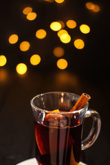 closeup pease of glass of mulled wine with orange and cinnamon on dark black background, on white plate, christmas lights, large yellow bokeh