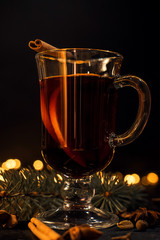 cinnamon lies in a glass, closeup glass of mulled wine with orange and cinnamon on dark black background,  Christmas tree and lights, large yellow bokeh, mulled wine set