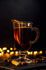 cinnamon lies in a glass, closeup glass of mulled wine with orange and cinnamon on dark black background,  Christmas tree and lights, large yellow bokeh, mulled wine set