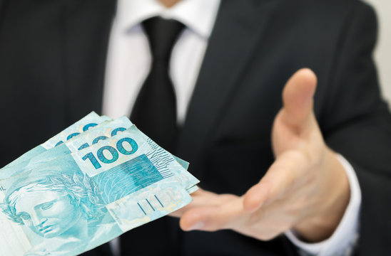 Close-up businessman wearing black suit and tie receiving Brazilian money notes. Brazil one hundred (100) reais. Selective focus.