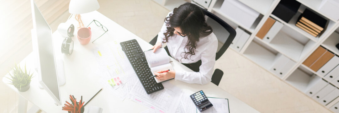 A young girl is sitting at the computer desk in the office, holding a pen in her hand and looking at the notepad.