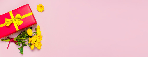 Red gift box with gold ribbon and yellow chrysanthemums on pink background top view flat lay. Holiday concept, birthday gift, valentine day or anniversary. Congratulations background copy space.