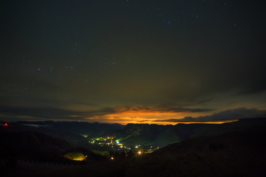 Bright stars shine through the clouds in the night sky over a city in a mountain valley. Overcast night in the summer.