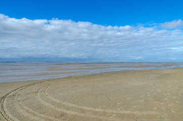 Fototapeta na wymiar Flat sandy beach with car tracks and footprints close to shallow water