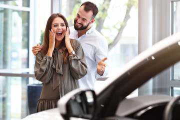 Adult man making surprise to beautiful woman in car showroom