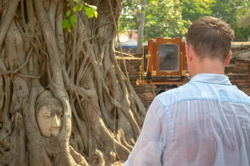 Photographer with retro camera with bellows accordion shooting Ayutthaya sights, Buddha head in...