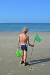 Junge von hinten, steht mit  K&auml;scher und Eimer am Sandstrand vor dem Meer mit blauem Himmel