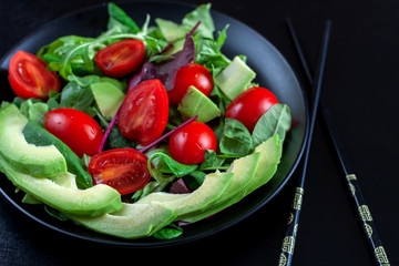 Vegetable salad with avocado, cherry tomatoes, arugula and spinach on a black plate on a dark background.