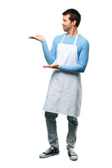 A full-length shot of a Man wearing an apron extending hands to the side and smiling for presenting and inviting to come on isolated background