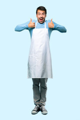 Full body of Man wearing an apron giving a thumbs up gesture and smiling because has had success on blue background