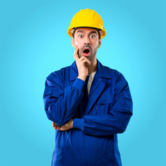 Young workman with helmet surprised and shocked while looking right. Expressive facial emotion on blue background