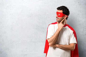 Superhero man with mask and red cape standing and looking to the side with the hand on the chin on textured grey background © luismolinero