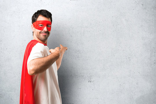 Superhero Man With Mask And Red Cape Pointing Back With The Index Finger Presenting A Product From Behind On Textured Grey Background