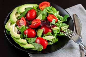 Vegetable salad with avocado, cherry tomatoes, arugula and spinach on a black plate on a dark background.