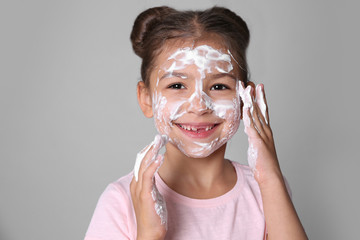 Cute little girl with soap foam on face against gray background